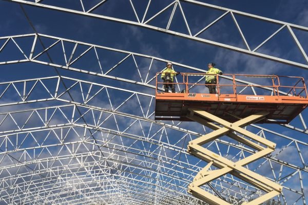 Two workers on a scissor lift at a construction site, framed by a metal structure against a blue sky.