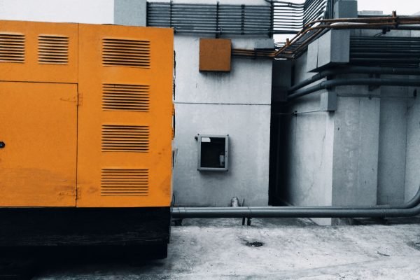Outdoor view of an industrial generator with visible piping and wiring against a concrete wall.