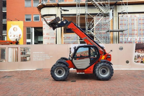 A red telehandler parked at a construction site with scaffolding and building materials in the background.